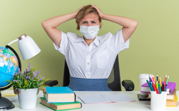 Frowning young blonde student girl wearing protective mask sitting at desk with school tools keeping hands on head looking at camera isolated on olive green wall