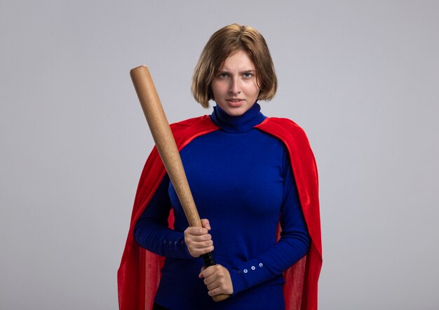 Frowning young blonde superhero girl in red cape holding baseball bat  isolated on white wall with copy space