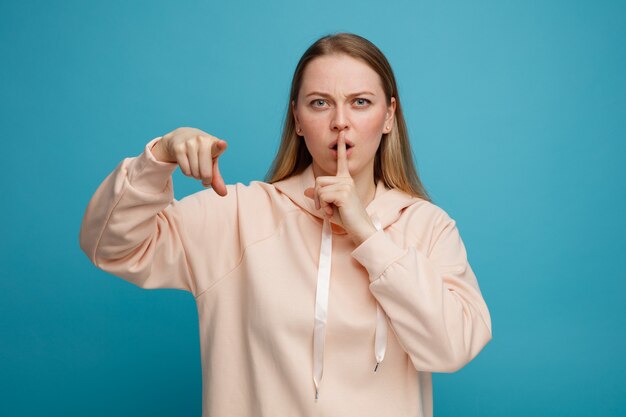 Frowning young blonde woman looking and pointing at camera doing silence gesture 