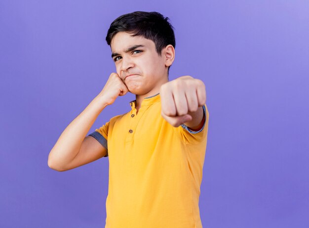 Frowning young caucasian boy  touching face with fist stretching out fist towards isolated on purple wall with copy space