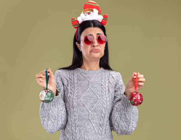 Frowning young caucasian girl wearing santa claus headband with glasses holding christmas baubles looking at camera isolated on olive green background