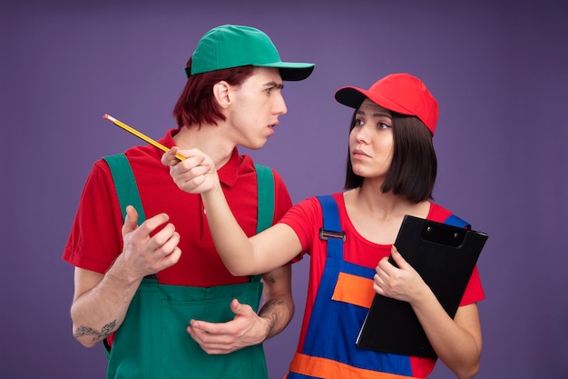 Free photo frowning young couple in construction worker uniform and cap girl holding pencil and clipboard looking and pointing at side with pencil guy keeping hand in air looking at girl isolated