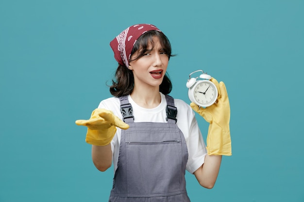 Frowning young female cleaner wearing uniform bandana and rubber gloves showing alarm clock looking at camera showing go away gesture isolated on blue background