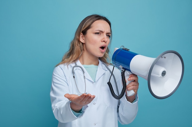 Frowning young female doctor wearing medical robe and stethoscope around neck looking at side pointing at side with hand talking by speaker 