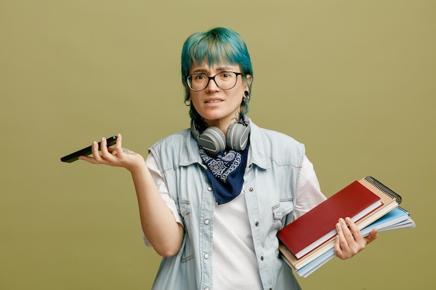 Free Photo frowning young female student wearing glasses bandana and headphones around neck holding note pads and mobile phone looking at camera isolated on olive green background