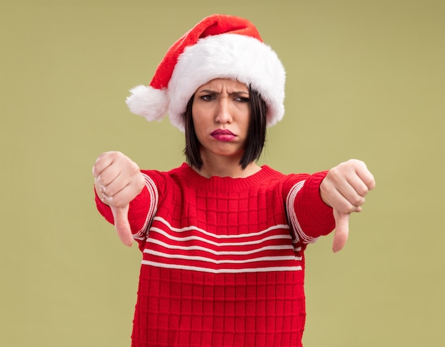 Frowning young girl wearing santa hat looking at side showing thumbs down isolated on olive green background