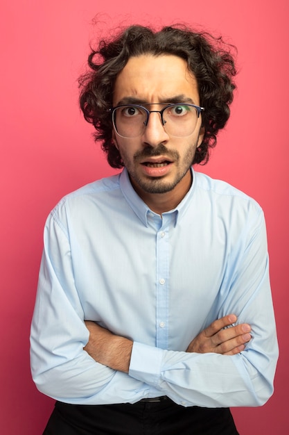 Frowning young handsome caucasian man wearing glasses sitting with closed posture  isolated on crimson wall