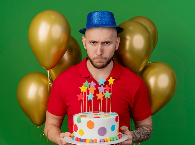 Frowning young handsome slavic party guy wearing party hat standing in front of balloons holding birthday cake looking at camera isolated on green background