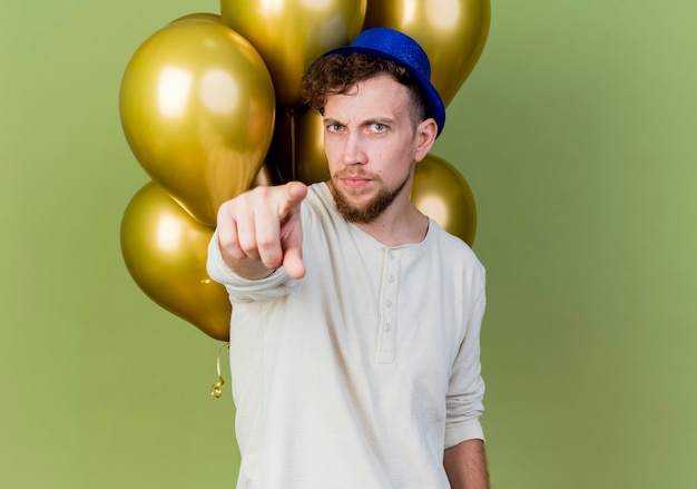 Frowning young handsome slavic party guy wearing party hat standing in front of balloons looking and pointing at camera isolated on olive green background with copy space