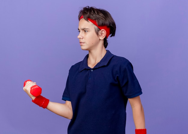 Free photo frowning young handsome sporty boy wearing headband and wristbands with dental braces looking at side holding dumbbell isolated on purple wall