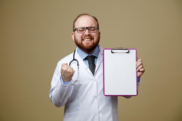 Free photo frowning young male doctor wearing glasses lab coat and stethoscope around his neck showing clipboard and yes gesture with eyes closed isolated on olive green background