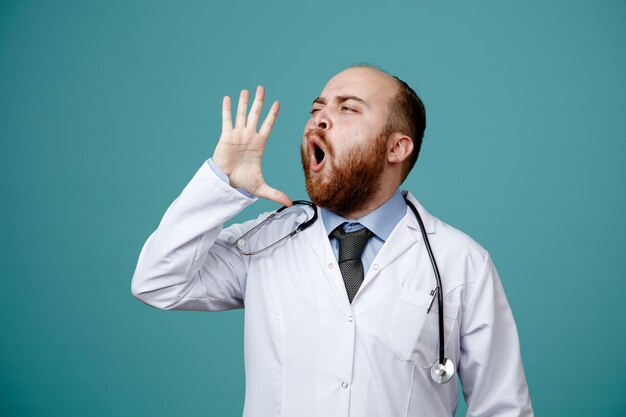 Frowning young male doctor wearing medical coat and stethoscope around his neck showing empty hand near head looking at side shouting something out isolated on blue background