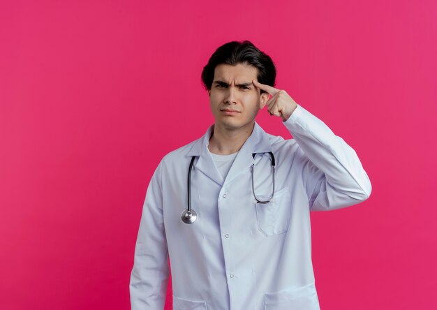 Frowning young male doctor wearing medical robe and stethoscope  pointing finger on temple isolated on pink wall with copy space