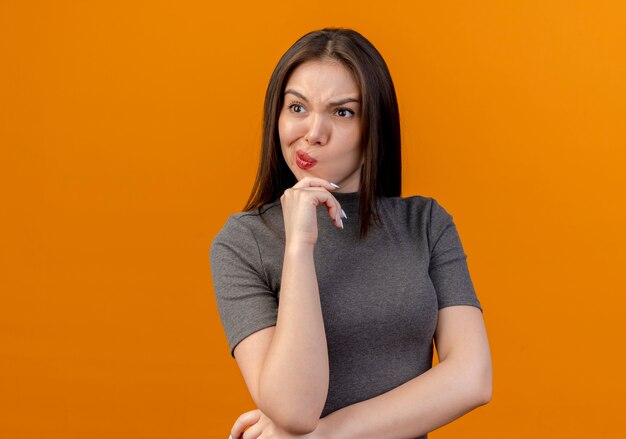 Frowning young pretty woman putting hands under elbow and on chin looking at side isolated on orange background with copy space