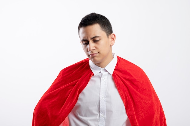 Frowning young superhero boy in red cape looking down isolated on white background