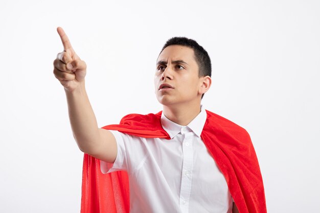 Frowning young superhero boy in red cape looking and pointing at side isolated on white background