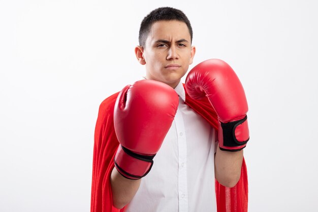 Frowning young superhero boy in red cape wearing box gloves looking at camera doing boxing gesture isolated on white background with copy space