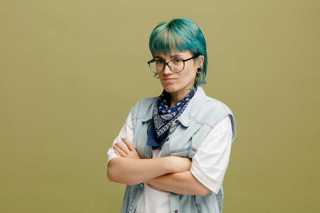 Free photo frowning young woman wearing glasses bandana on neck standing in profile view keeping arms crossed looking at camera isolated on olive green background