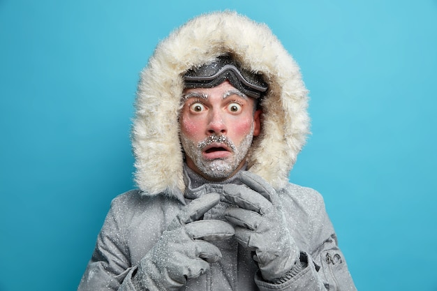 Frozen male explorer has red face covered with frost stares very shocked surprised by very low temperature wears warm jacket and gloves has walk outdoor during blizzard cold weather