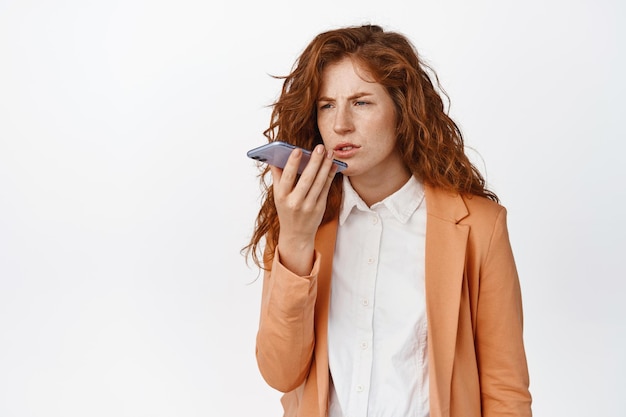 Free photo frustrated businesswoman recording voice message translating on mobile phone app with confused face expression standing against white background