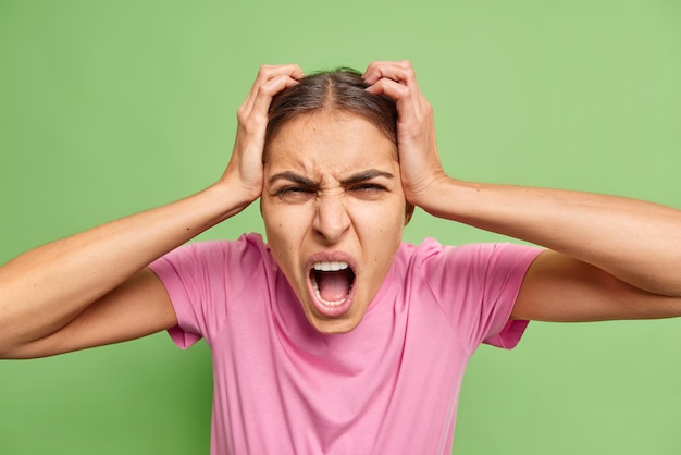 Free Photo frustrated crazy woman keeps hands on head yells loudly from despair feels irritated stands bothered and pressured loses control has menntal breakdown poses against green wall