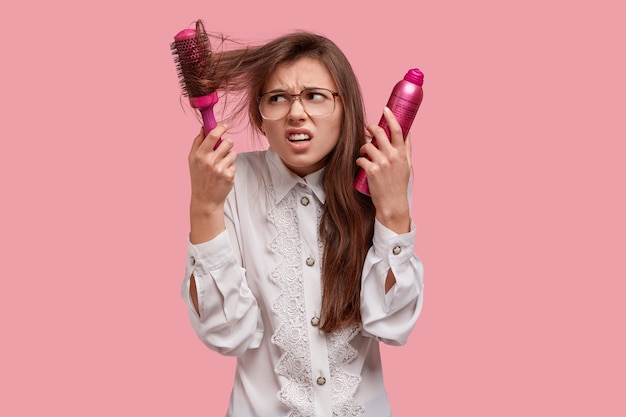 Free photo frustrated discontent woman tries to blow hairbrush out of hair, holds hairspray, has problem with hair, dressed in white shirt frowns face with displeasure isolated on pink wall. beauty concept