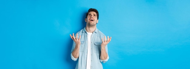 Free photo frustrated guy complaining to god looking up and pleading standing against blue background upset