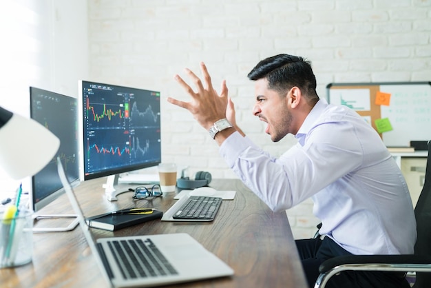 Free photo frustrated hispanic young freelance stock trader shouting while looking at graphs on computer screen