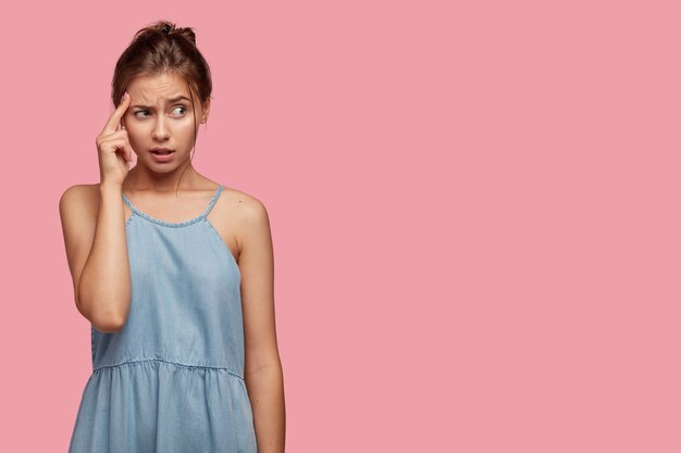 Frustrated student keeps finger on temple, frowns face in displeasure, tries to find solution in mind, ponders how solve problem, wears denim dress, models over pink wall with copy space aside