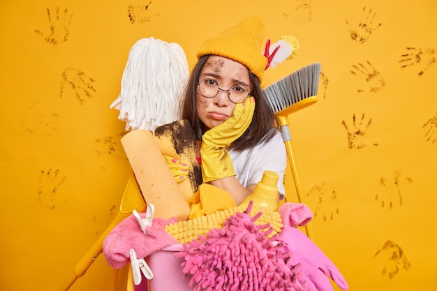 Free photo frustrated tired asian girl doesnt want to clean up room surrounded by lots of cleaning tools and detergents looks sadly at camera isolated over yellow wall