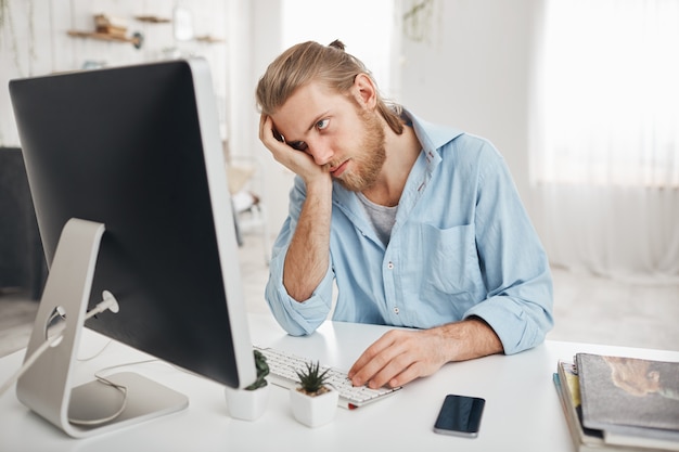 Frustrated tired bearded Caucasian employee touching his head, feeling absolutely exhausted because of overwork, calculating accounts, sitting in front of computer screen. Deadline and overwork