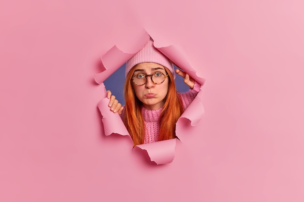 Free photo frustrated unhappy redhead young european woman purses lower lips wears hat and knitted jumper looks sadly through paper