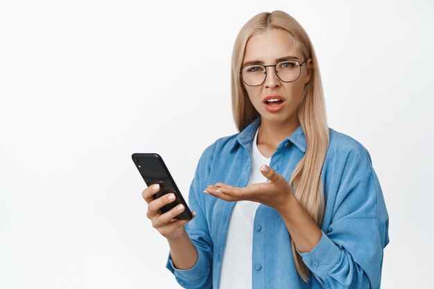 Frustrated woman in glasses complaining at smartphone, holding mobile phone and looking puzzled on white.