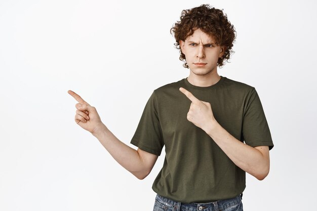 Frustrated young man with curly hair pointing and looking left with angry face expression standing in tshirt over white background