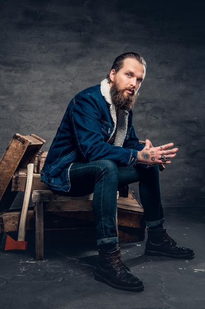 Free photo full body portrait of bearded hipster male wearing a denim jacket sits on a wooden box.