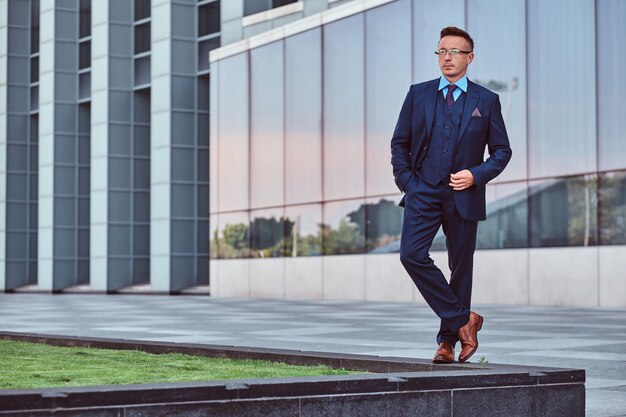 Full body portrait of a confident man in an elegant suit posing with hand in pocket against a skyscraper background.