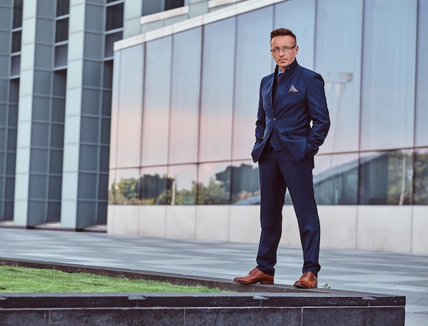 Free Photo full body portrait of a confident man in elegant suit standing with hands in pockets against a skyscraper background.