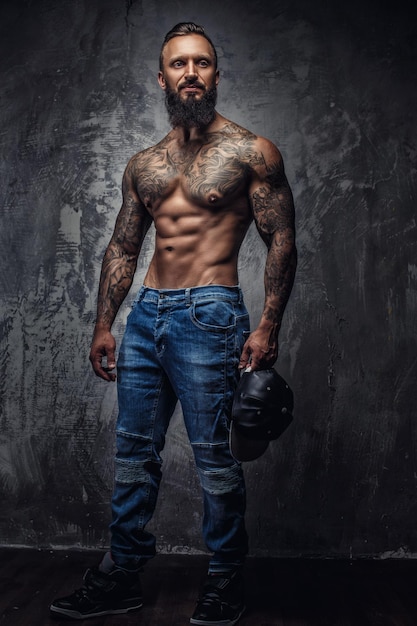 Full body portrait of muscular tattooed bearded man in blue jeans over grey wall.