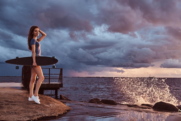 Free photo full body portrait of a sensual girl holds a skateboard while standing on the beach is enjoying amazing dark cloudy weather during sunset.