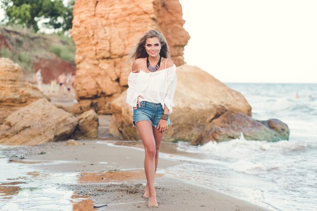 Full-lengh photo of pretty blonde girl with long hair  walking on the beach near sea. She is smiling to the camera.