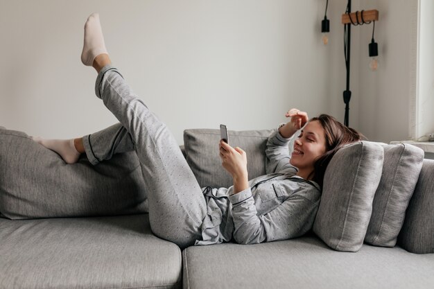 Full-lenght indoor shot of happy woman in home clothes scrolling smartphone while lying on the coach in sunny day.