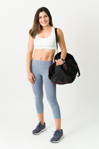 Free photo full length of an attractive female athlete wearing sportswear smiling while carrying a gym bag. confident young woman with toned abs