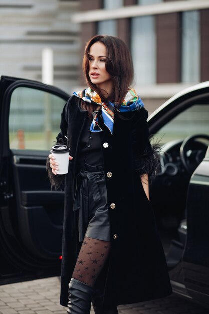 Full length of a fashionable lady in black coat and leather boots wearing colorful handkerchief around her neck. She is carrying a cup of takeaway coffee against open car in the street.