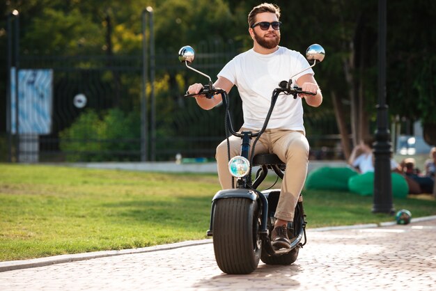 Full length of happy bearded man in sunglasses rides on modern motorbike outdoors and looking away