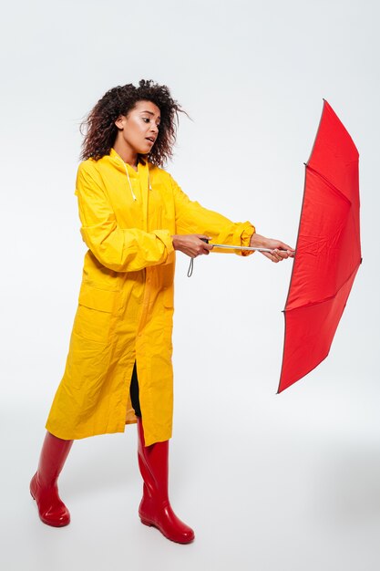 Full-length image of confused african woman in raincoat opening umbrella