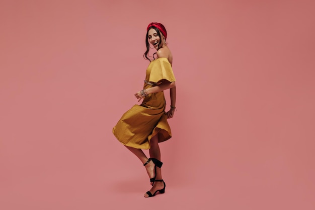 Free photo full length photo of cheerful woman in midi yellow dress and black stylish heels dancing and looking into camera on pink backdrop