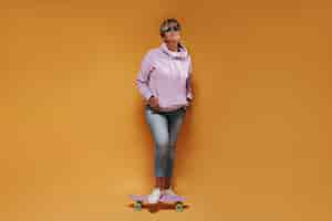 Free photo full length photo of cool woman with short hair in sunglasses, wide hoodie and skinny jeans smiling and posing with pink skateboard.