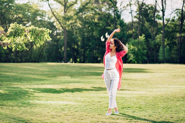 Full-length photo of cute brunette girl walking in summer park. She wears white clothes,  long pink shirt. She is enjoying.