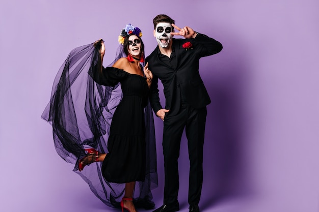 Full-length photo of funny guy and girl in masquerade masks laughing and posing in good mood. Lady in black veil with flowers in her hair touches boyfriend showing peace sign.