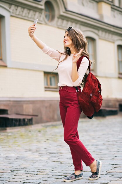 Full-length photo of pretty girl with long hair making selfie-portrait on phone in city. She has vinous color on clothes and looks enjoyed.